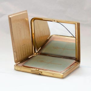 May include: A gold-toned compact mirror with a hinged lid and a small mirror inside. The compact has a light green interior with the text "After Shave" printed on it.