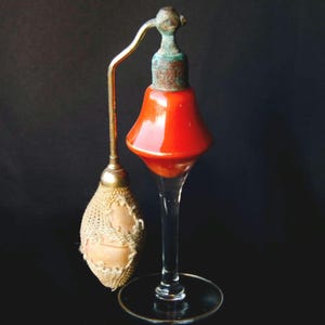 Può includere: Un vaporizzatore di profumo vintage in vetro con un bulbo in vetro rosso e un ugello spray in metallo dorato. Il vaporizzatore ha una base in vetro trasparente con un bordo in metallo dorato.