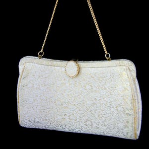 Könnte beinhalten: Eine weiße Clutch mit einer goldenen Kettenkette und einem goldenen Verschluss. Die Clutch hat ein weißes und goldenes Brokatmuster.