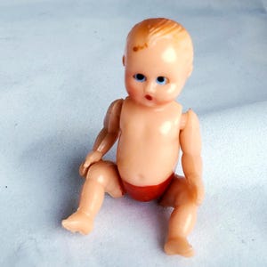 May include: A small plastic baby doll with blue eyes and a red nappy. The doll is sitting with its legs crossed and its arms outstretched.