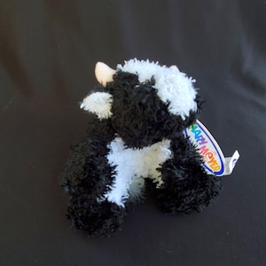 May include: A small, plush cow toy with black and white fur. The toy has a pink nose and a tag that reads "Mary Meyer". The cow is sitting on a black surface.