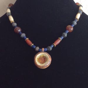 May include: A necklace with a round pendant made of brown and gold ceramic. The necklace is made of various colored beads, including red, blue, and white.