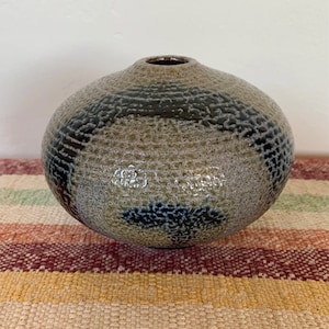 May include: A small, round, ceramic vase with a textured, speckled glaze in shades of gray, brown, and blue. The vase has a wide opening at the top and a rounded bottom.