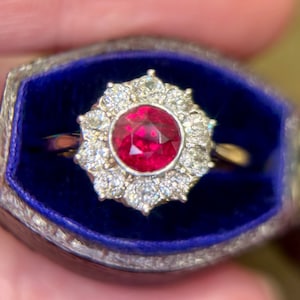 May include: A ring with a large red ruby surrounded by a halo of smaller diamonds. The ring is in a blue velvet ring box.