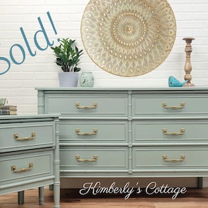 May include: A light green bamboo dresser with six drawers and gold hardware. A smaller matching dresser with two drawers is to the left. The dressers are in front of a white brick wall. The text "Sold!" is written in blue on a white background in the top left corner of the image.