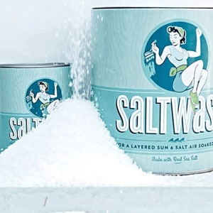 May include: Two blue and white cans of salt wash, one larger than the other, with a retro-style illustration of a woman on each. The larger can has the text "SALTWASH" and "FOR A LAYERED SUN & SALT AIR SOAKED LOOK" and "Made with Real Sea Salt". The smaller can has the text "Salt". White salt is spilling out of the larger can.