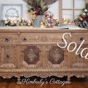 May include: A vintage wooden buffet with intricate carvings and a distressed finish. The buffet has six drawers and two doors, and it is sitting on a wooden floor. The word "Sold!" is written in white on the front of the buffet.