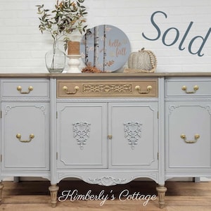 May include: A gray painted vintage buffet with gold hardware and a wood top. The buffet has four doors and three drawers. The top drawer is a different color than the rest of the buffet. The buffet is sitting on a white brick wall background. The words "Sold" are written in gray on a white background.