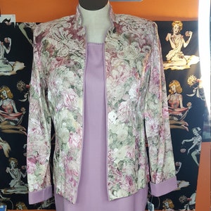 Puede incluir: Un vestido lavanda con una chaqueta a juego con estampado floral. La chaqueta presenta un fondo crema con estampados florales rosas y verdes. El vestido tiene un diseño sencillo y elegante.