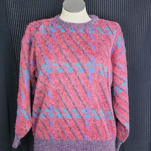 May include: A vintage sweater with a vibrant pattern. The sweater features a red base with diagonal stripes in black, accented by blue and purple geometric shapes. The collar, cuffs, and hem are a solid purple color. The sweater is displayed on a mannequin.