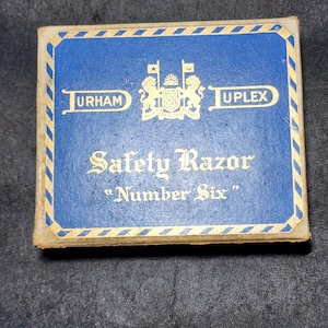 Może przedstawiać: Zabytkowe pudełko na maszynkę do golenia Durham-Duplex. Pudełko jest niebieskie ze złotymi obramowaniami i napisami. Tekst na pudełku brzmi "Durham Duplex Safety Razor Number Six". Pudełko ma złoty herb pośrodku.