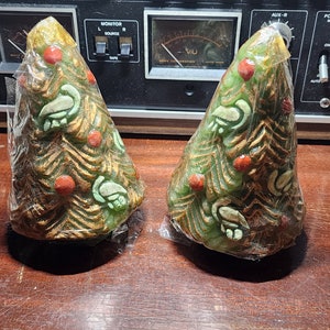 May include: Two green, gold, and red Christmas tree candles wrapped in plastic. The candles are decorated with gold swirls and red berries.