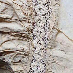 May include: Close-up of a beige fabric with a white crocheted trim. The fabric is gathered and has a slightly wrinkled texture.
