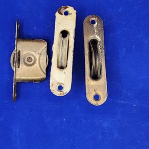 May include: Three vintage metal pulleys and a latch on a blue surface. The pulleys are rectangular with a central wheel and mounting holes. One pulley is painted white, showing signs of wear. The latch is a small, rectangular box with a wheel.