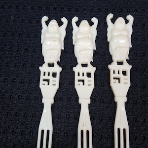 May include: Three white plastic forks with a decorative design of a person with raised arms. The forks are on a black background.