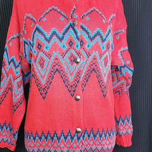 May include: A red cardigan sweater with a geometric pattern in navy blue and light blue. The sweater has a round neckline, long sleeves, and silver-colored buttons down the front. The pattern includes zigzags and diamond shapes.