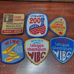 Pode incluir: Seis patches bordados com pinos de boliche e o texto "WIBC League Champion" para diferentes anos. Os patches são azuis, amarelos, vermelhos, brancos e marrons.