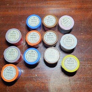 May include: Twelve small, round, colorful containers with white labels that read "NEWS LESSON" and include the volume and date. The containers are arranged in a circle on a wooden surface.