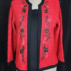 May include: A red open-front cardigan with black trim and floral embroidery. The cardigan features a textured knit fabric and is displayed on a mannequin. The embroidery includes black flowers and silver sequins.
