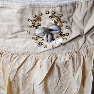 May include: A cream-colored skirt with a gathered ruffle at the bottom. The skirt has a decorative embellishment of gold sequins and white seashells.