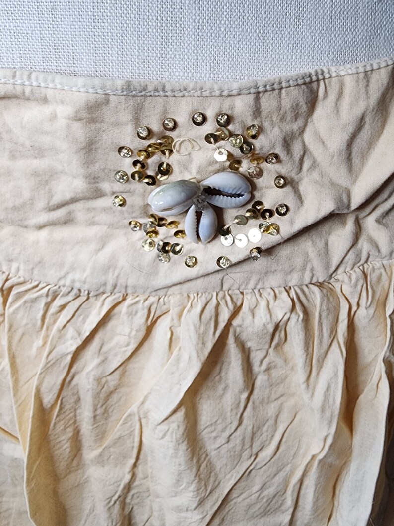 May include: A cream-colored skirt with a gathered ruffle at the bottom. The skirt has a decorative embellishment of gold sequins and white seashells.
