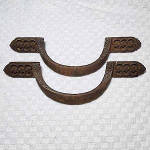 May include: Two matching semi-circular metal drawer pulls with a dark, weathered bronze finish. Each handle features a textured, ornate pattern on the mounting plates and a smooth, curved grip section, showcasing a distinct aged patina throughout.