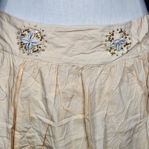 May include: A cream-colored skirt with a gathered waist and two decorative embellishments made of seashells and gold sequins.
