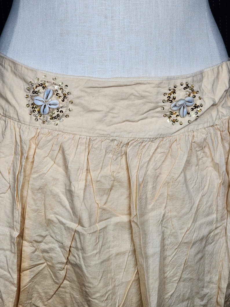 May include: A cream-colored skirt with a gathered waist and two decorative embellishments made of seashells and gold sequins.