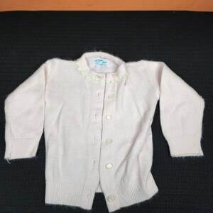 May include: A pale pink knitted cardigan with a white lace trim around the neckline. The cardigan has a button-up front and long sleeves.