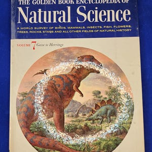 Puede incluir: Un libro antiguo de tapa dura titulado "The Golden Book Encyclopedia of Natural Science". La portada presenta una gran ilustración de dinosaurios en un paisaje prehistórico. El título del libro está en letras grandes y azules.