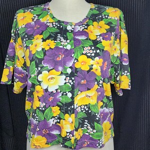 May include: A short-sleeved button-up shirt with a vibrant floral pattern. The shirt features a black background with large yellow and purple flowers, green leaves, and small white flower clusters. The shirt has a round neckline and white buttons.