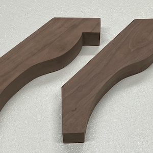 May include: Two wooden corbels with a dark brown finish. The corbels have a curved design with a rectangular top and bottom. The wood grain is visible. These are likely for home decor or construction.