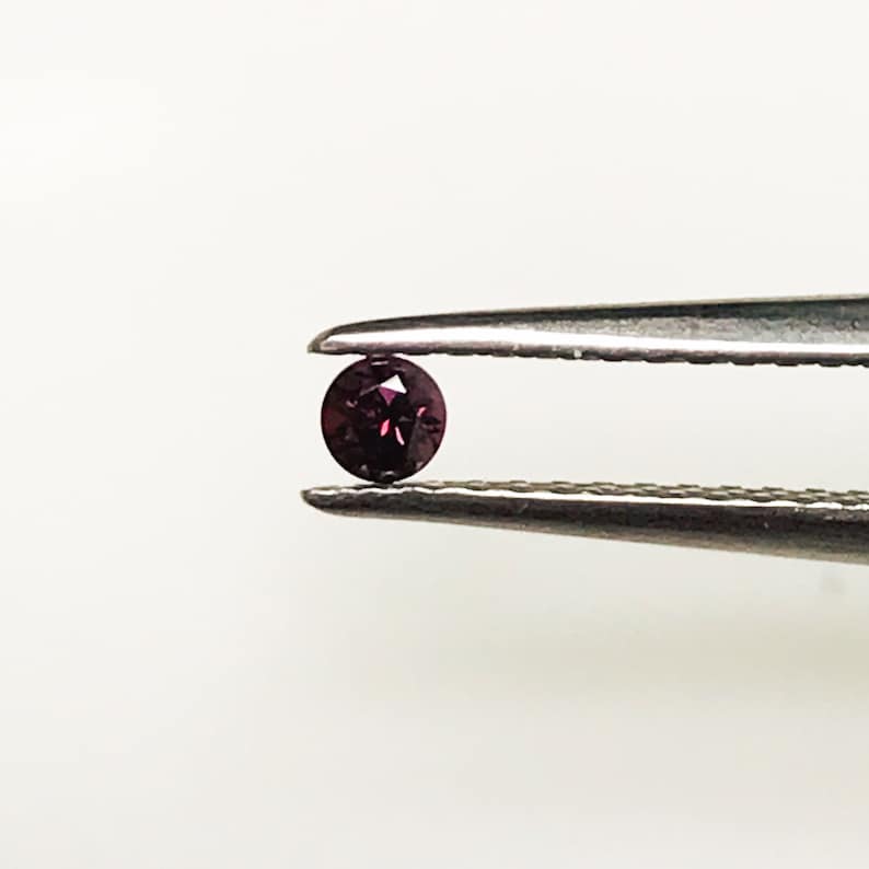 May include: A small, round, dark purple gemstone held in tweezers.