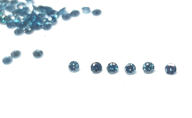 May include: A collection of small, round, faceted gemstones in a deep teal blue colour. Six individual stones are arranged in a horizontal line, while a larger cluster of identical loose stones is scattered in the upper left corner of the frame.