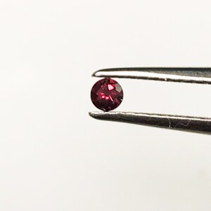 May include: A small, round, red gemstone held in tweezers. The gemstone is a deep red color and has a slight sparkle.
