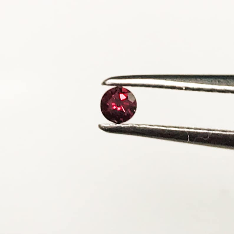 May include: A small, round, red gemstone held in tweezers. The gemstone is a deep red color and has a slight sparkle.