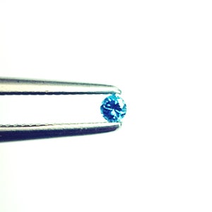 May include: A small, round, brilliant-cut gemstone with a vibrant blue colour held between the tips of a pair of silver-toned metal tweezers. The gemstone features multiple facets that reflect light, creating a sparkling appearance against a plain background.