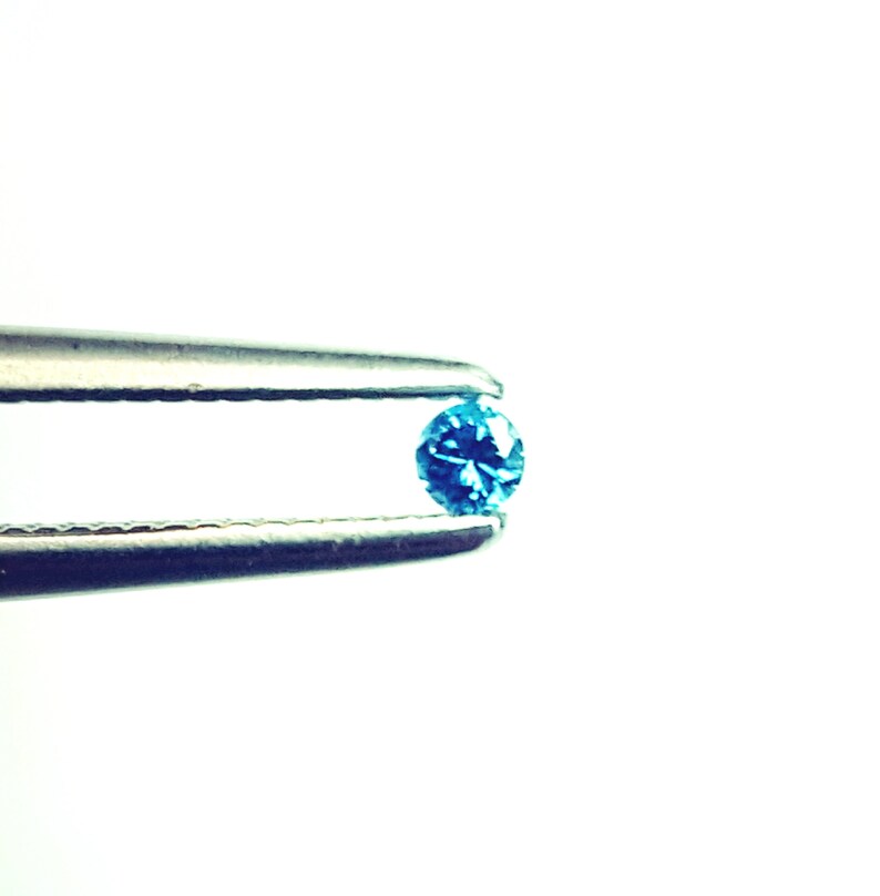 May include: A small, round, brilliant-cut gemstone with a vibrant blue colour held between the tips of a pair of silver-toned metal tweezers. The gemstone features multiple facets that reflect light, creating a sparkling appearance against a plain background.