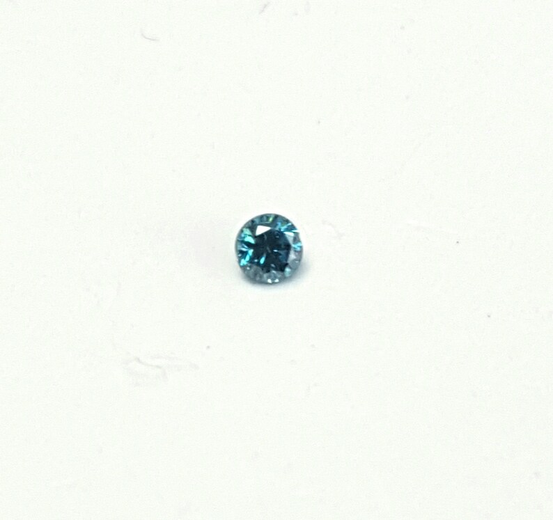 May include: A small, round, faceted gemstone with a vibrant teal-blue colour. The stone features a brilliant cut with multiple reflective surfaces that catch the light, creating a sparkling appearance against a plain, neutral white background.