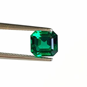 Puede incluir: Una esmeralda verde vibrante con un corte cuadrado, sostenida por unas pinzas sobre un fondo blanco.
