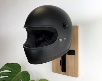 May include: A black motorcycle helmet with a matte finish is mounted on a wooden wall hook. The hook is made of metal and has a simple design.