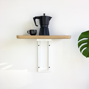 May include: A white shelf bracket with a wooden shelf, a black coffee pot, and a black coffee cup on top. The shelf is mounted to a white wall.