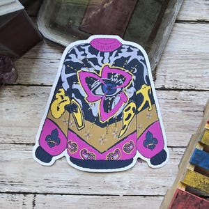 May include: A sticker in the shape of a sweater with a pink, yellow, and blue design. The sweater has a pink collar and cuffs, with a large pink and blue floral design on the front. The sleeves are yellow and blue, with a fleur-de-lis and paisley pattern.