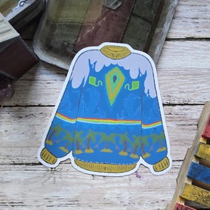 May include: A sticker featuring a blue sweater design with a white top, yellow collar and cuffs, and a rainbow stripe. The sweater has a central green and orange diamond shape and palm trees along the bottom.