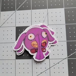 May include: A pink plush toy with stitched edges, large eyes, and a stitched mouth. The toy has a red bow tie and a white flower on its chest.