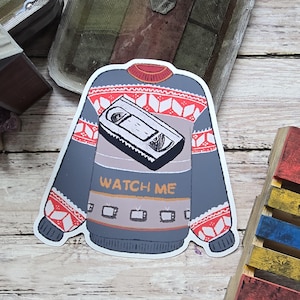 May include: A sticker in the shape of a sweater with a VHS tape design. The sweater is gray with red and white geometric patterns on the sleeves and shoulders. The text "WATCH ME" is printed on the front, with small TV icons below.