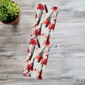 May include: A white bookmark with a repeating pattern of knives and red blood splatters.