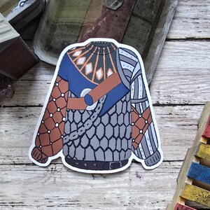 May include: A sticker featuring a stylized illustration of a sweater. The design incorporates shades of blue, brown, gray, and white, with geometric and chain-like patterns. The sweater has a collar, sleeves, and a belt.