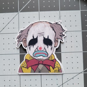 May include: A sticker of a sad clown with a red bow tie, yellow jacket, and blue and red makeup. The clown has a white face with black eyes and a blue mouth.