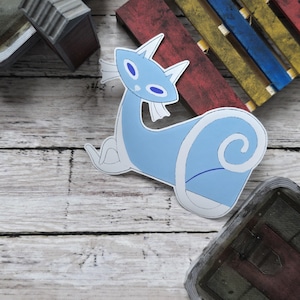 May include: A light blue sticker featuring a stylized cat-like creature with white accents. The sticker has a curved body and a long neck, with pointed ears and oval eyes. The sticker is on a wooden surface.
